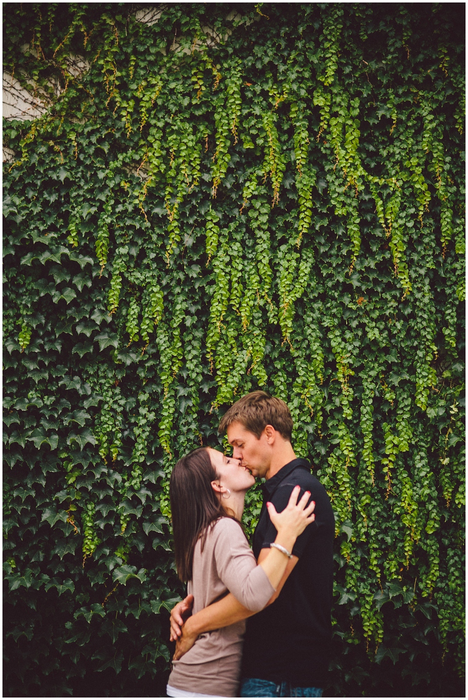 engaged couple kissing in front of green ivy wall