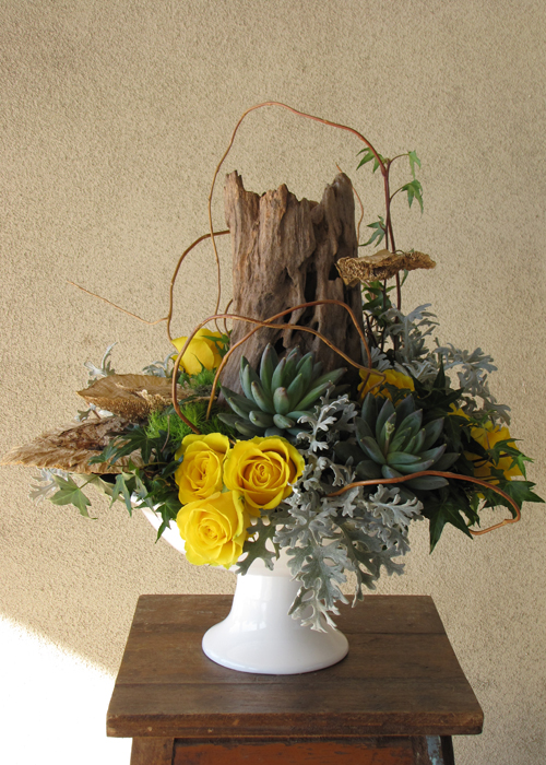 woodland arrangement with a reclaimed wood stump, sponge mushrooms, curly willow tips, yellow roses, Green Trick carnations, ivy,  Pachyveria succulents, and dusty miller in a ceramic pedestal bowl