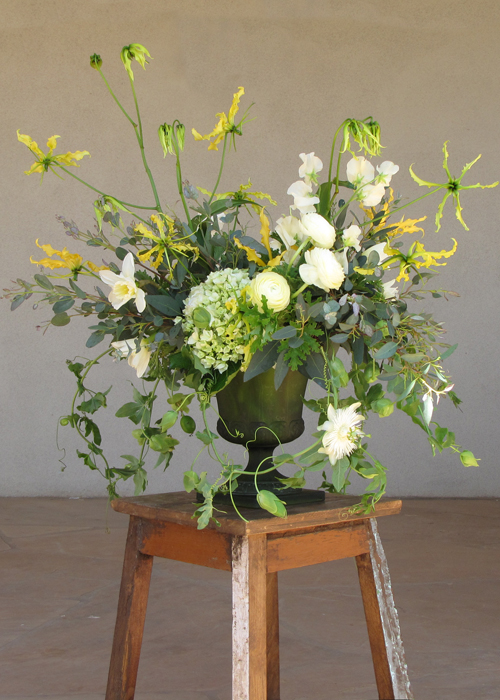 centerpiece with ivory sweet peas, ivory ranunculus, ivory daffodils, yellow gloriosa lily, green hydrangea, scented geranium, gunnii eucalyptus, and passion vine, arranged in a vintage green glass urn