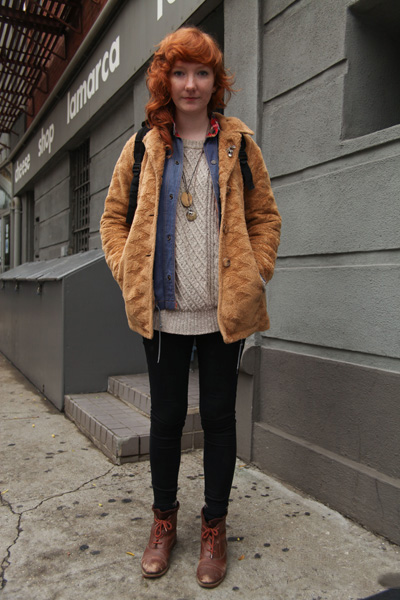 School of Visual Arts SVA in New York City student wears a faux fur tawny jacket over a blue denim shirt with plaid collar detailing, worn open over a chunky crew neck cream off white sweater and black leggings tucked into brown boots with red laces. All of this was accesorized with a silver fleur de lis lapel pin, a bronze leaf pendant, a silver stag necklace and a mountain print brown corduroy two tone backpack.