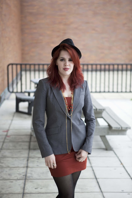 Red haired Lucy, an Advertising Photography major at the Rochester Institute of Technology RIT wears a black fedora and a grey blazer over a short cropped red dress and golden red jewelry.